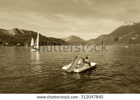 ANNECY LAKE, FRANCE - AUGUST 27, 2015: Tourists enjoy water sports (pedaling, sailing, kayaking) at Annecy lake surrounded by beautiful mountains. Annecy Lake is one of most popular French resorts.