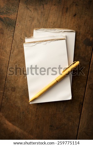 Blank memo pad and gold pen on old grungy wooden surface with dim lighting. Intentionally shot and with low key shadows