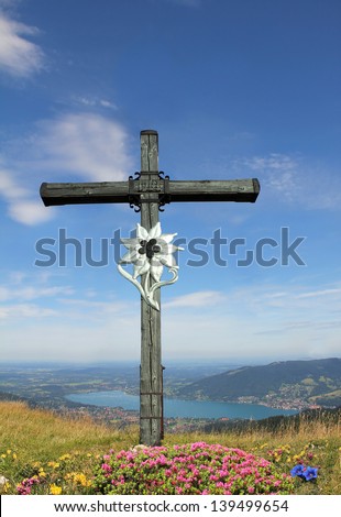 wooden mountain cross with edelweiss and alpine flowers, view to bavarian lake tegernsee , german landscape