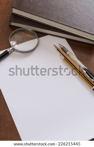Pen and pencils on blank paper with magnifying glass and stack of books.
