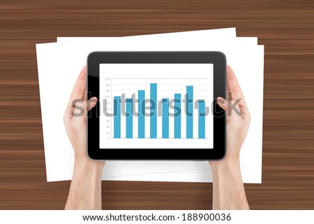 Hand holding digital tablet with bar chart graphic and white, blank note sheet on wooden background.