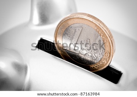 Close-up full frame image of one Euro coin partially inserted into the slot of a silver piggy bank.