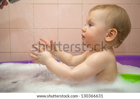 Small baby girl with red hair washes in a baby bath with water spray and bubbles