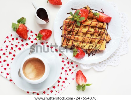 Belgium waffles with strawberries and chocolate decoration on plate with cup of coffee and small jar with chocolate on white table, selective focus