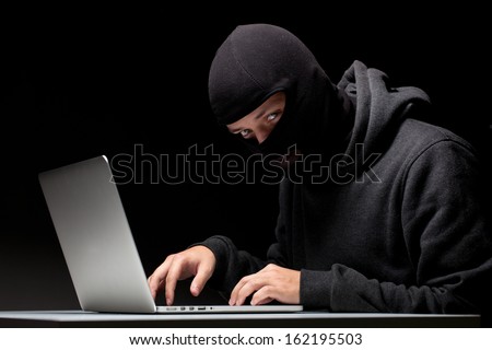 Computer hacker in a balaclava working in the darkness stealing data and personal identity information off a laptop computer