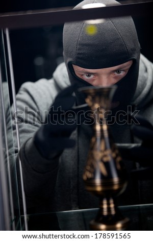 Angry burglar wearing black woollen mask, considering vase over black