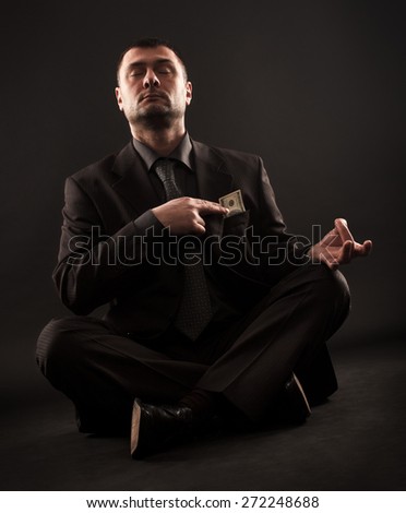Businessman sitting in lotus position doing yoga and holding one hundred dollar bill on his heart with right hand.