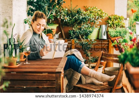 Young beautiful woman relaxing on cozy balcony, reading a book, wearing warm knitted pullover, glass of wine on wooden table