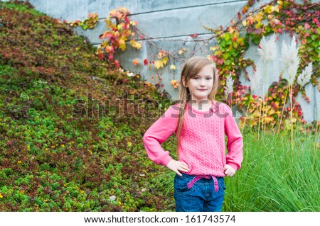 Outdoor portrait of a cute little girl in a garden on a nice autumn day, wearing grey coat, jeans, pink pullover and brown boots