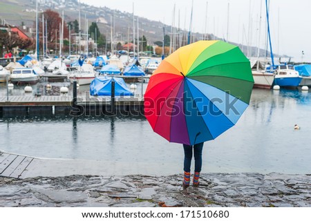 Child under big colorful umbrella watching the rain on the lake, view from the back