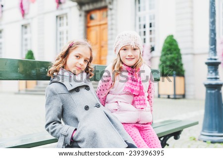 Outdoor portrait of two adorable little girls of seven years old, resting on the bench in a city