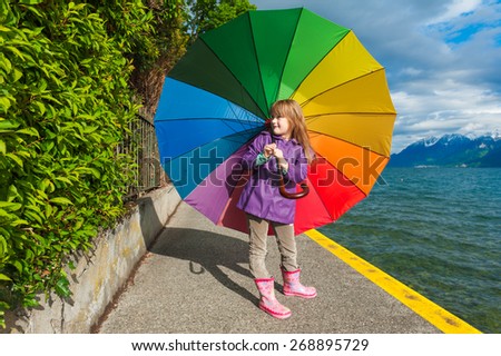 Outdoor portrait of a cute little girl with big colorful umbrella, wearing purple rain coat, boots, standing next to lake