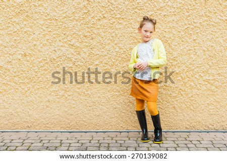 Fashion portrait of a cute little girl of 7 years old, wearing blouse, yellow jacket, orange skirt and black rain boots