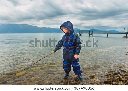 Cute little boy playing by the lake, pretending fishing, wearing blue waterproof all-in-one suit and rain boots