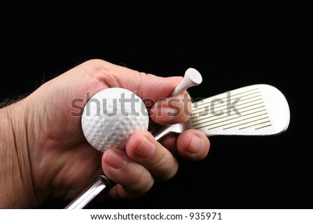 Hand holding golf club, ball & tee with black background.