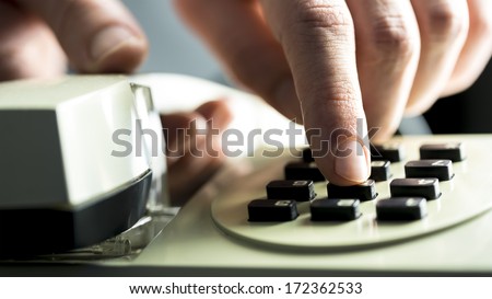 Man dialing on a landline telephone instrument entering the numbers on the buttons on the keypad while holding the handset, close up view