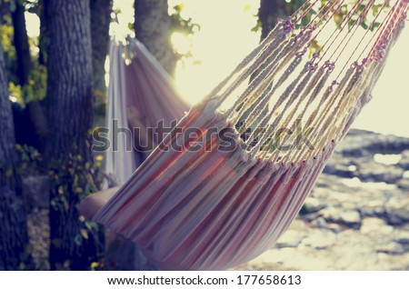 Retro effect faded and toned image of a person relaxing in a hammock in the shade of a tree on a shoreline viewed from behind.