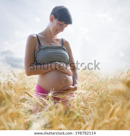 Cheerful young woman in late pregnancy standing in the middle of golden wheat field looking lovingly at her swollen belly and enjoying her life while bonding with the unborn child.