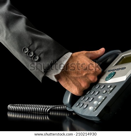 Close-up of the hand of a businessman picking up the receiver of a black land line telephone, placed on the desk, with copy space on black