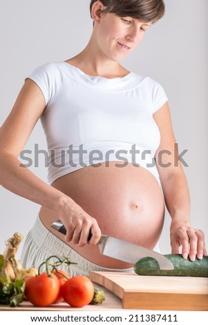 Attractive pregnant young woman with a bare swollen baby belly standing at a kitchen counter making a healthy salad chopping a cucumber and tomatoes with a quiet smile of anticipation.