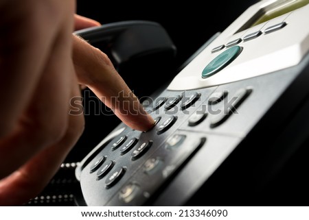 Person dialing out on a landline telephone punching in the numbers on the keypad with a finger, closeup side angle view on a dark background.