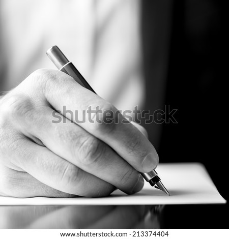 Low angle black and white image of a male hand holding a fountain pen as though writing on a blank sheet of paper with shallow dof in square format.