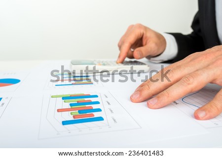 Close up view of the hands of a businessman analyzing a bar graph using a manual calculator to check the statistics and projections.
