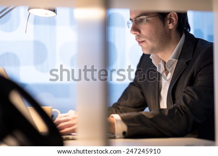 Attractive young businessman wearing glasses sitting working at his desk typing information into a laptop computer with a mug of coffee close at hand, side view.