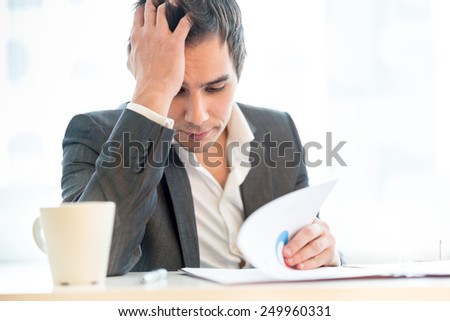 Tired diligent hardworking businessman resting his head on his hand as he reads through a lengthy report with a mug of energizing coffee alongside him, close up view of him sitting at his desk