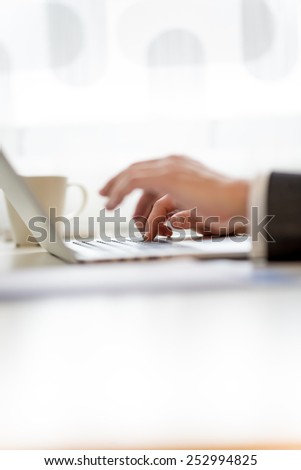 Man typing in information on his laptop with selective focus to the hand furthest from the camera, side view of both hands on the desk.