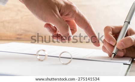 Male hand pointing to a divorce paper on which a woman is busy writing with a pen in a close up conceptual view.