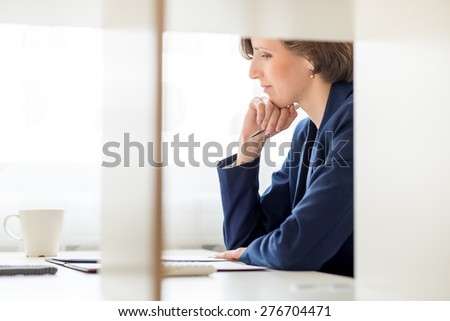 Businesswoman sitting reading a document at her desk with an engrossed expression, side view through an interior office partition.