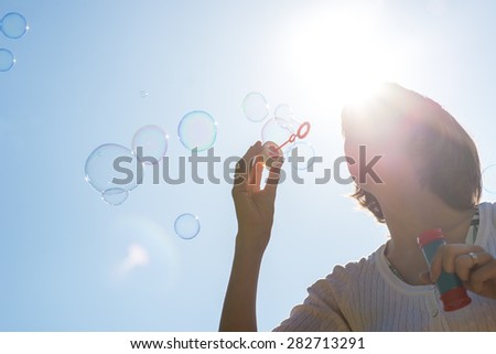 Low angle close up view of a young woman against the sun in a clear blue sky joyfully blowing a stream of soap bubbles.