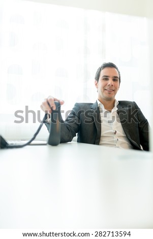 Smiling Young Businessman at his Office Holding Telephone on Top of his Desk While Looking at the Camera.