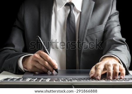 Front view of male graphic designer or photographer  in an elegant suit sitting at his office desk working with stylus pen on digital tablet and simultaneously using computer keyboard.