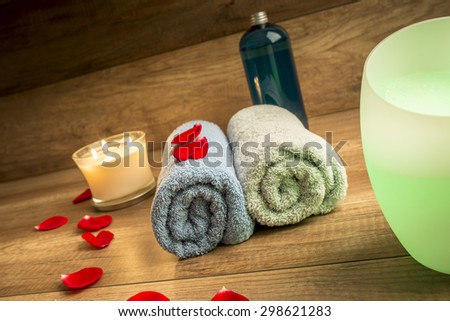 Romantic Spa Still Life Concept for a Couple with Two Rolled Towels, Lit Candle, Bottle of Massage Oil and Essential Oil Diffuser Surrounded by Red Rose Petals with Wooden Background.