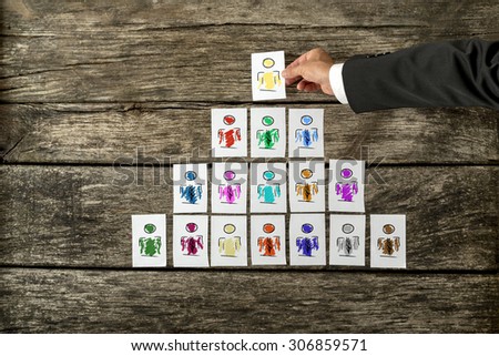Leadership and team management concept with a businessman arranging a series of hand-drawn cards depicting people into a pyramid and just about to place the last card at the pinnacle in place.