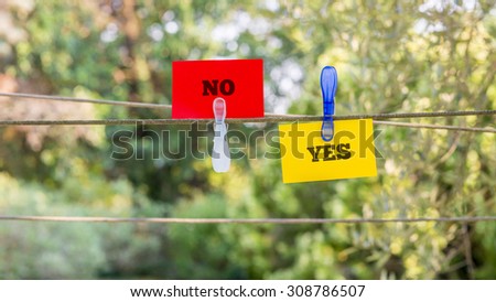 Conceptual Red and Yellow Paper with Yes and No Messages Clipped on a String Against Fuzzy Greenery Background.
