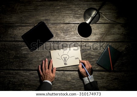 Overhead view of a businessman working late at his table by the light of a desk lamp creating new ideas with light bulb drawn on paper, digital tablet and note with pen also lying on his wooden desk.