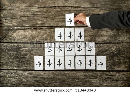 Overhead view of banker or financial adviser arranging white cards with Dollar sign in a pyramid shape on textured wooden desk.