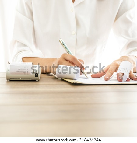Front view of female bank employee in elegant white blouse writing something on receipts with documents and adding machine on her wooden desk.
