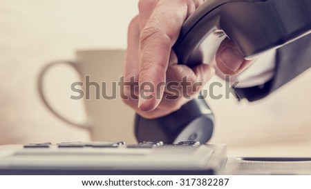 Retro image of male hand dialing a telephone number on a black phone with cup of coffee in background.