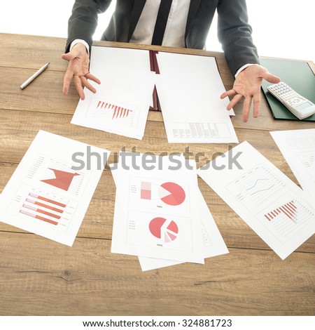 Busy financial adviser working at his business desk with many documents, charts and reports placed all over the table, showing a doubtful gesture with his hands.