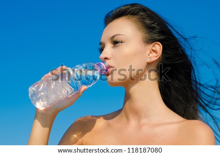 Beautiful brunette girl, with long hair in the wind, drinking from a bottle of water. Outdoors, blue sky background.