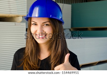 Beautiful girl wearing a blue safety helmet, happy worker signing thumbs up. Construction site background.