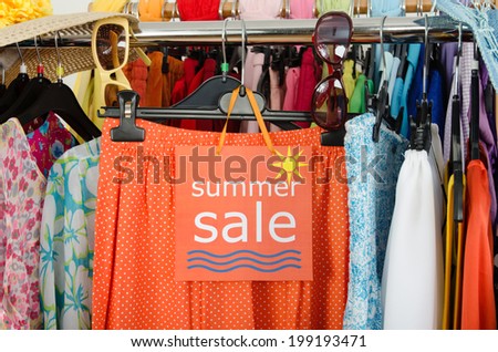 Close up on a big sale sign for summer clothes. Clearance rack with colorful summer outfits and accessories displayed on hangers.