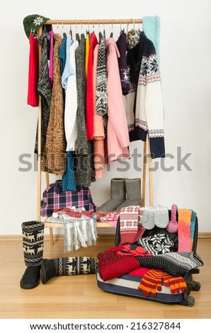 Packing the suitcase for winter vacation. Wardrobe with clothes nicely arranged and a full luggage. Dressing closet with colorful winter clothes and accessories on hangers.