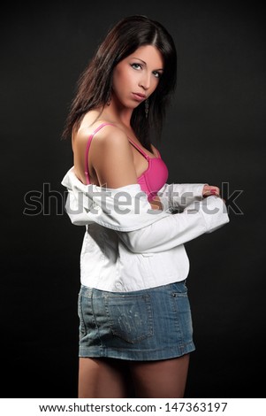 Beautiful brunette in a white ladies blazer and pink bra and denim skirt posing in a studio, on a black background, fashion and glamour photography