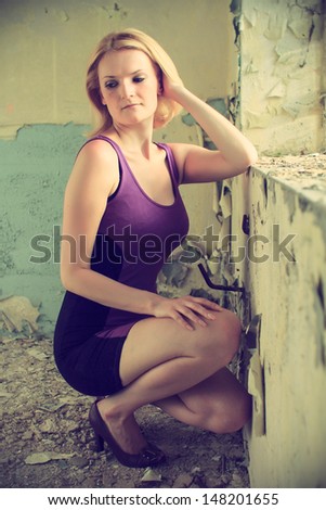 Beautiful blond woman in a purple dress posing in a old building, she kneels at the window, cross processing, fashion photography