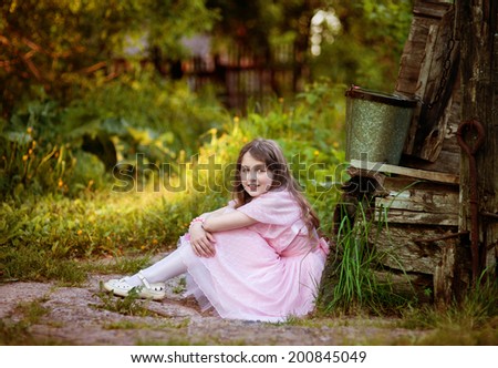 The nine-year-old girl in a pink dress sits in a summer garden at an old well in a sunny day